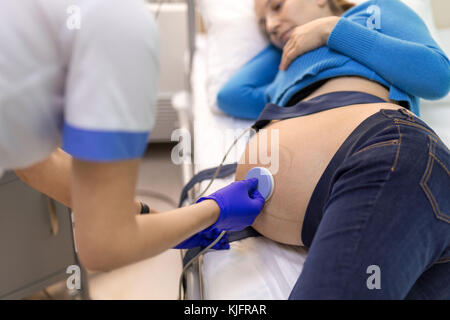 Giovane donna felice sdraiato su un letto in un ospedale medico mentre l'impostazione di un sensore la cardiotocografia per esaminare il feto heartbeat. Foto Stock