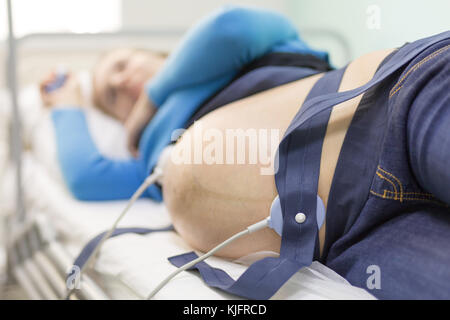 Giovane donna felice sdraiato su un letto in un ospedale medico mentre l'impostazione di un sensore la cardiotocografia per esaminare il feto heartbeat. Foto Stock