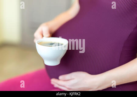 Giovane donna incinta seduta in una cucina e tenendo un bicchiere od un caffè in una mano. Pro e contro il concetto di caffè. Foto Stock