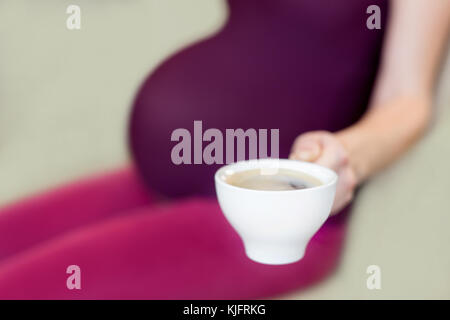 Giovane donna incinta seduta in una cucina e tenendo un bicchiere od un caffè in una mano. Pro e contro il concetto di caffè. Foto Stock