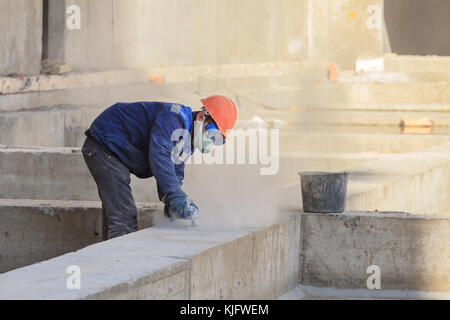 Il lavoratore in tuta e una maschera protettiva macina la parete in cemento. Foto Stock
