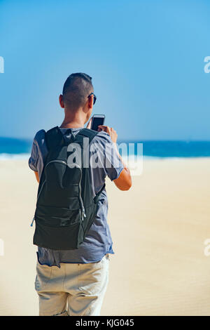 Primo piano di un giovane uomo caucasico portando uno zaino utilizzando uno smartphone all'aperto, con l'oceano sullo sfondo Foto Stock