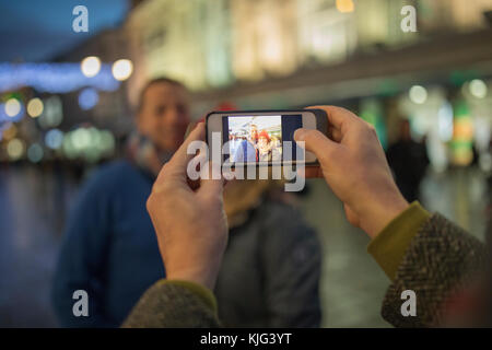 Punto di vista colpo di un telefono intelligente che viene utilizzato per prendere una foto di adulti nella festosa centro città a tempo di Natale. Foto Stock