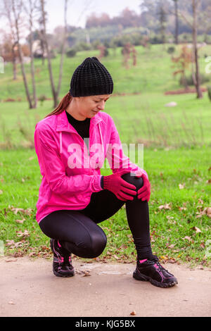 Atleta femminile male da un infortunio al ginocchio in una fredda giornata invernale sulla via della formazione di un parco urbano. giovane donna indossando giacca a vento rosa, beanie, g Foto Stock