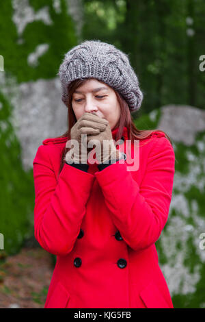 Giovane donna i brividi di freddo e di soffiaggio di aria calda per le mani su una foresta indossando un rosso lungo cappotto o soprabito, un beanie e dei guanti di protezione durante il periodo invernale Foto Stock