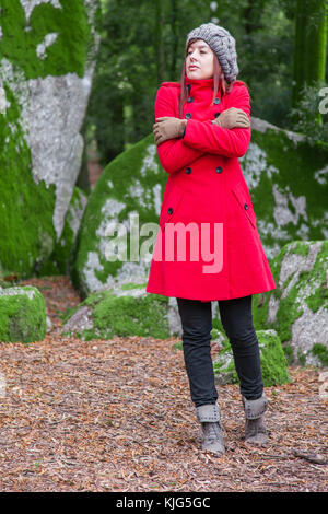 Giovane donna perso su una foresta i brividi di freddo e abbracciando o azienda stessa, indossando un rosso lungo cappotto o soprabito, un beanie e guanti durante la fal Foto Stock