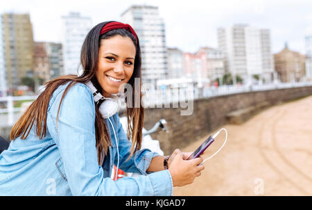 Spagna Gijon, sorridente ragazza con un telefono cellulare e le cuffie a waterfront promenade Foto Stock