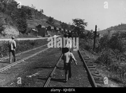 Una fotografia di diverse persone a piedi lungo i binari del treno, alcuni uomini e un ragazzo sono a piedi verso la città al fine di rilievo ricevuto cibo essendo fornito dalla Farm Security Administration, la fsa è stato creato come parte del new deal sotto il presidente franklin d roosevelt per fornire assistenza ai poveri agricoltori e le loro famiglie in tutto il paese la parte della città che è visibile è costituito da strutture identiche costruito sul fianco di una collina vicino alla stazione del treno che viaggia attraverso la città, Scott's run, West Virginia, 1935. dalla biblioteca pubblica di new york. Foto Stock