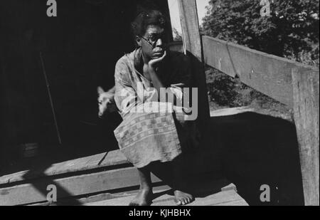 Una fotografia di una donna che indossa un semplice abito a fantasia, è seduta sul suo portico di legno mentre attende un incontro con un agente di soccorso dell'Agenzia di servizi agricoli, la FSA stava lavorando all'epoca per migliorare la vita degli agricoltori in tutto il paese, il suo cane sta uscendo dalla casa dietro di lei, Scott's Run, West Virginia, 1935. Dalla New York Public Library. Foto Stock