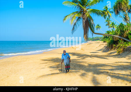 Bentota, Sri lanka - 6 dicembre 2016: l'anziano pescatore a piedi con un bastone lungo la spiaggia paradisiaca con palme e brillanti acque blu dell india Foto Stock