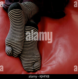 Due piedi in bianco e nero a strisce calze, incrociati a caviglie, appoggiato sulla pelle di colore rosso cuscino Foto Stock