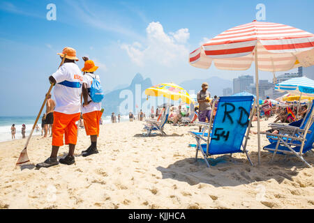 Rio de Janeiro - marzo 06, 2016: spiaggia pulitori su occupato Ipanema beach con ombrelloni e sedie con due fratelli montagne sullo sfondo. Foto Stock
