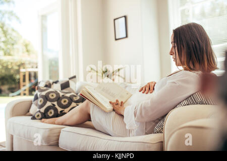 donna incinta seduta sul divano toccando la pancia e leggendo un libro. Aspettarsi che la madre si rilassasse sul divano preparasse un libro. Foto Stock