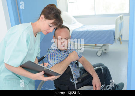 Uomo in sedia a rotelle in ospedale con un infermiere con clipboard Foto Stock