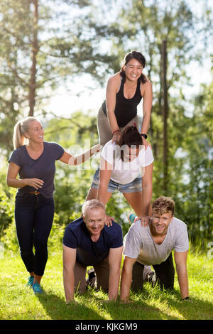 Sorridente maschio e femmina collaboratori rendendo piramide umana sul campo erboso in foresta Foto Stock