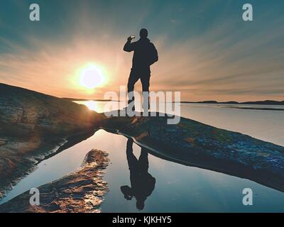 Silhouette posteriore di viaggio uomo prendendo selfie in mare. turistico con zaino in piedi su una roccia che si affaccia su un mare azzurro e prendendo la foto dal suo smart Foto Stock