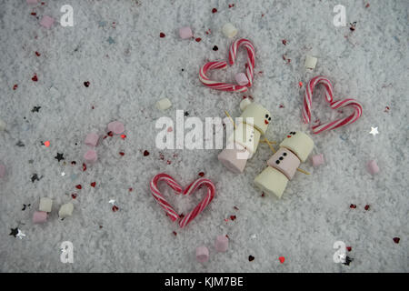 Romantico Inverno immagine fotografia di marshmallows a forma di pupazzo di neve con iced su sorrisi e giacente nella neve con cuori rossi stelle e candy canes Foto Stock