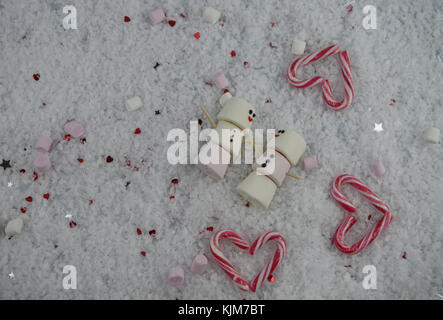 Romantico Inverno immagine fotografia di marshmallows a forma di pupazzo di neve con iced su sorrisi e giacente nella neve con cuori rossi stelle e candy canes Foto Stock