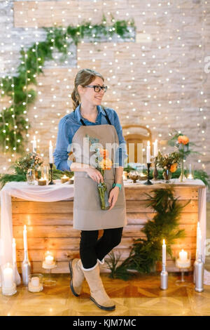 Design floreale, professione, concetto di decorazione. carina ragazza sorridente vestito con grembiule pulito e confortevole shirt azienda Vaso con fiori arancione raccolti nel piccolo ma grazioso mazzetto su sfondo di festa Foto Stock