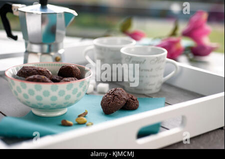 Rustico cacao Biscotti e pistacchi sul vassoio in legno con caffettiera, tazze, pistacchi freschi, cubetti di zucchero e fiori in background Foto Stock