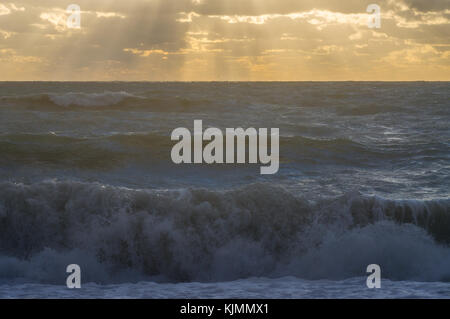 Un enorme ondata cade sulla riva e i raggi del sole attraverso le nuvole di illuminare la tempesta sul mare Foto Stock