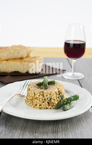 Risotto con asparagi con la piastra di ceramica su tavolo in legno con argento forcella. in background sale di due pezzi di focaccia e un bicchiere di vino rosso Foto Stock