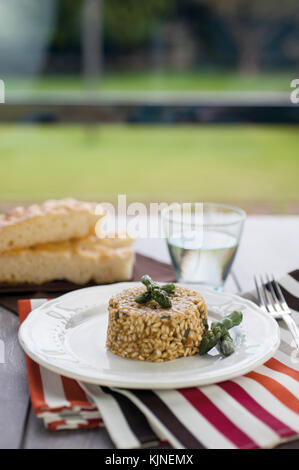 Risotto con asparagi con la piastra di ceramica su tavolo in legno con argento forcella, bicchiere di acqua e a strisce colorate tovaglia. in background due s Foto Stock