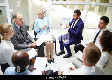 Gruppo di professionisti seduti in cerchio e prendendo parte alla discussione di nuove strategie di lavoro Foto Stock