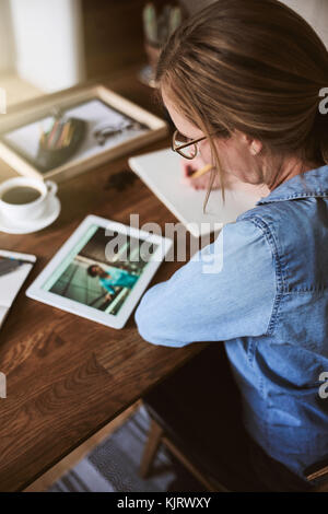 Giovane donna seduta da sola a un tavolo di lavoro con una tavoletta digitale e scrivere le idee su un blocco note Foto Stock