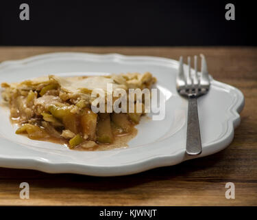 Una fetta di torta di mele sulla piastra bianca sul tavolo di legno, visto dal lato, decorata con una forcella, contro la parete nera Foto Stock