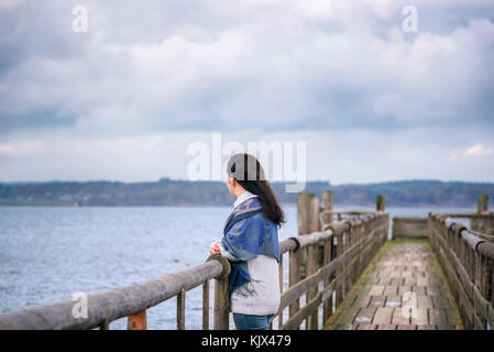 Giovane donna in piedi da solo, su un vecchio ponte di legno, di pensare e di ammirare l'orizzonte nel pomeriggio, oltre il lago di Chiemsee, in Baviera, Germania. Foto Stock