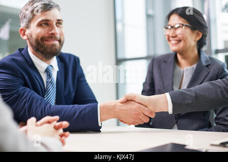 Ritratto di fiduciosa imprenditore seduto al tavolo della sala riunioni moderna e agitando la mano di un irriconoscibile business partner come segno di successo Foto Stock