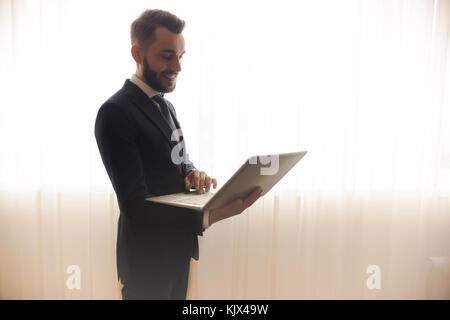 Vista laterale del successo imprenditore barbuto utilizzando laptop e sorridente in piedi contro lo sfondo della finestra tende in camera di albergo, spazio di copia Foto Stock