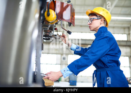 Occupato con esperienza operaio di fabbrica in hardhat concentrandosi sul lavoro e mediante macchina perforatrice in officina Foto Stock