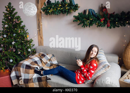 Felice ragazza guardare contenuti in streaming su linea in uno smart phone seduto su un divano in inverno a casa Foto Stock