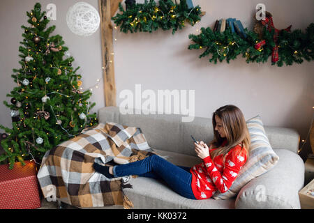 Felice ragazza guardare contenuti in streaming su linea in uno smart phone seduto su un divano in inverno a casa Foto Stock