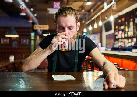 Vista anteriore verticale del moderno uomo barbuto ubriacarsi da soli in pub seduta a tavola e di bere whisky Foto Stock
