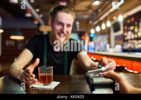 Ritratto di barbuto uomo ubriaco di pagare tramite smartphone acquistare bevande nel bar, concentrarsi sul terminale di pagamento in primo piano Foto Stock