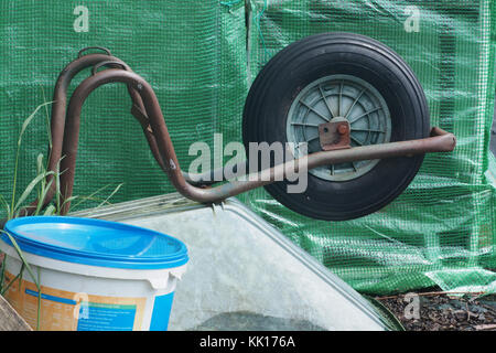 Ruota di una carriola rovesciata nella parte anteriore di una serra di plastica con una vasca di fertilizzante Foto Stock