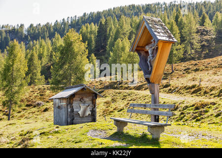 Antico crocifisso ligneo, banco e wc su un altopiano di pascolo con paesaggio alpino e montano in Carinzia occidentale, Austria. Foto Stock