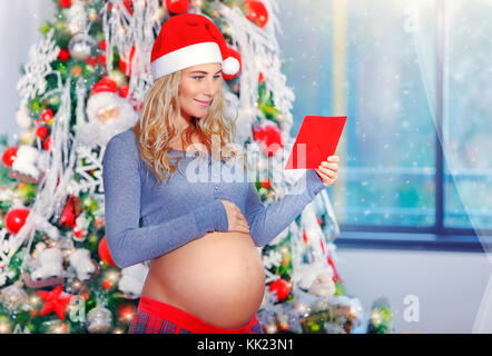 Felice donna incinta nella notte di natale di lettura la lettera, indossando red santa hat con piacere celebrare la vacanza invernale a casa in atmosfera accogliente Foto Stock