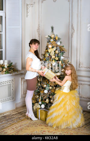 Mom rende la figlia di un regalo di Natale di circa decorate a nuovo anno albero. famiglia Capodanno e Natale. ragazza vestita a festa su una vacanza in famiglia Foto Stock