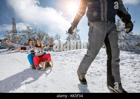 Felice ragazzo e ragazza seduta su slittini mentre il padre li trazione sulla neve Foto Stock