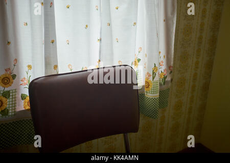 Imbottitura schienale di una sedia da cucina di fronte ad una bella tenda floreale con fiori di colore giallo in un ombroso interno Foto Stock