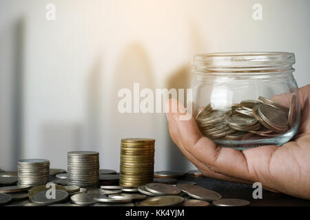 Mano che tiene il vaso di risparmio di denaro con la pila di monete - primo piano Foto Stock