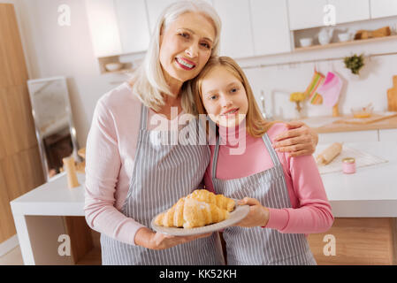 Fatto in casa. bel lieto abile la nonna e la nipote in piedi insieme e sorridenti durante la foratura di una piastra con croissant Foto Stock