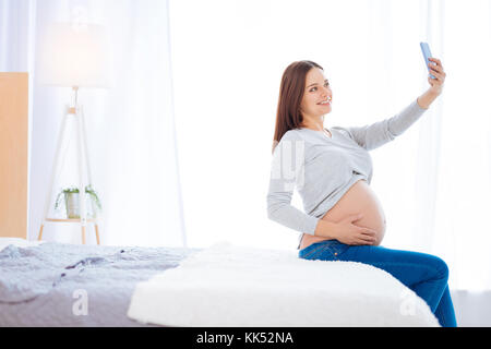 Bella foto. esuberante sorprendente carino donna incinta prendendo selfie mentre la pancia di levigatura con una mano e sedersi sul bordo del letto Foto Stock