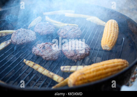 Patties di carne fresca e pannocchie di granoturco grigliare su un barbecue portatile su un viaggio di campeggio in una vista ravvicinata Foto Stock