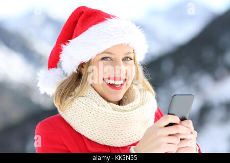 Donna felice guardando a voi in possesso di telefono nel natale con una montagna innevata in background Foto Stock