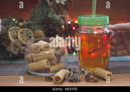 Mano che tiene un vetro chiaro tazza di tè con un cappuccio, una ciotola di cookies, una ghirlanda di Natale Foto Stock